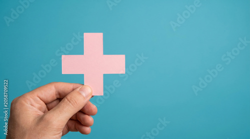 Human hand holding a soft pink plus sign cutout against a bright solid blue background