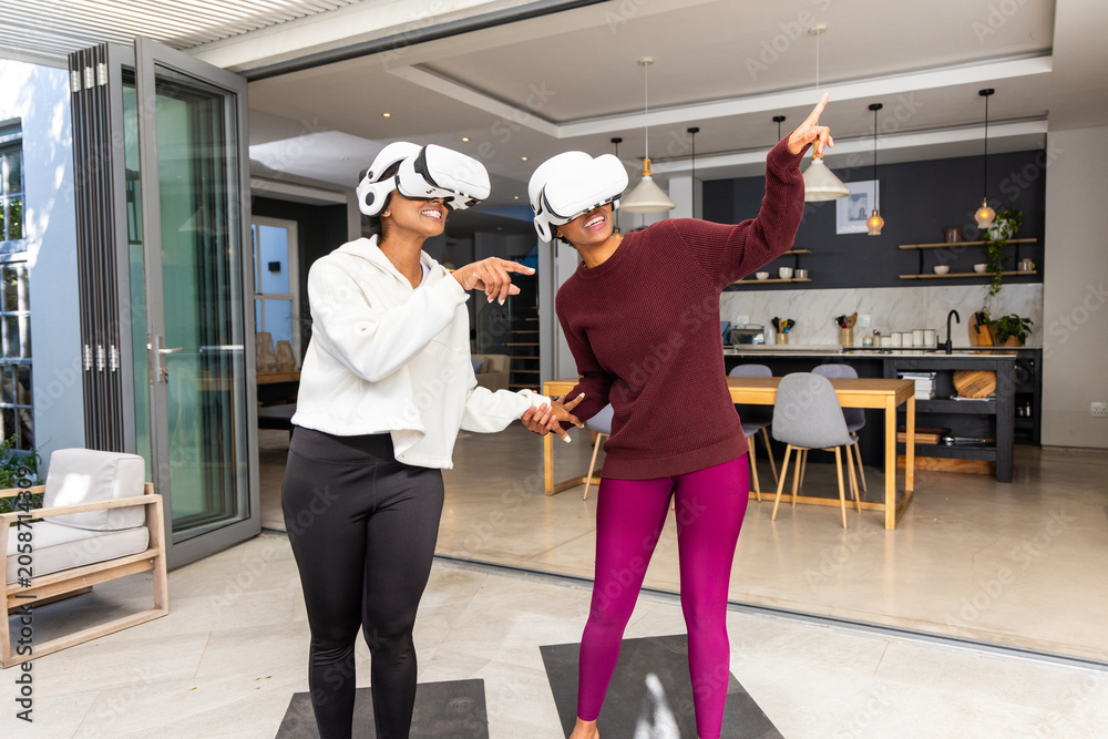 Obraz premium Diverse female friends holding hands and using white VR headsets on black mats near glass doors