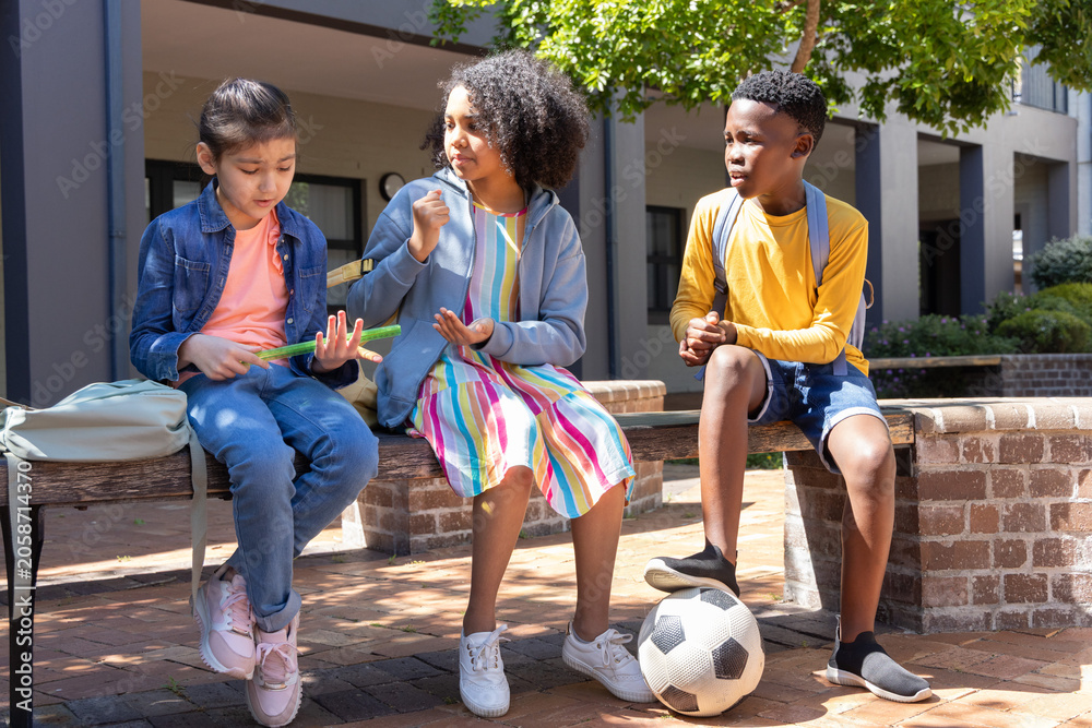 Naklejka premium Diverse school kids sitting on bench in brick courtyard, examining green snack, foot on soccer ball