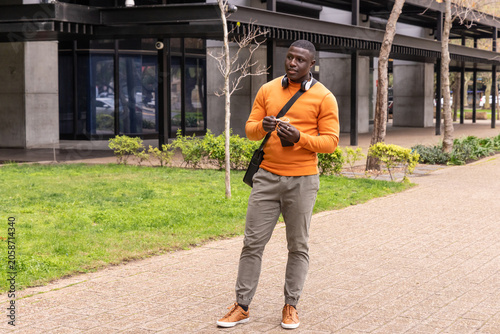 African American man standing on paved path holding keys wearing orange sweater and headphones