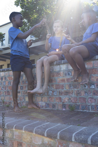 Three school-age children wearing blue shirts sitting on brick wall at schoolyard, holding snack