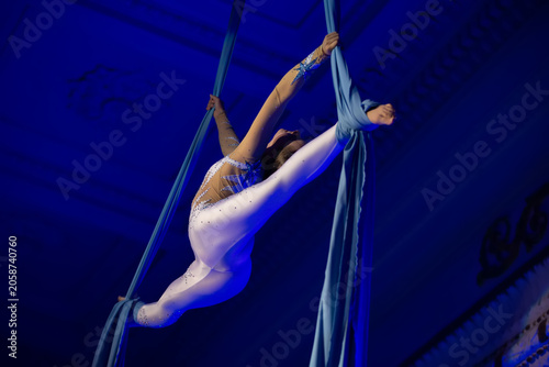 A gymnast demonstrates her aerial circus skills during an evening show with blue lighting and silk ribbons.