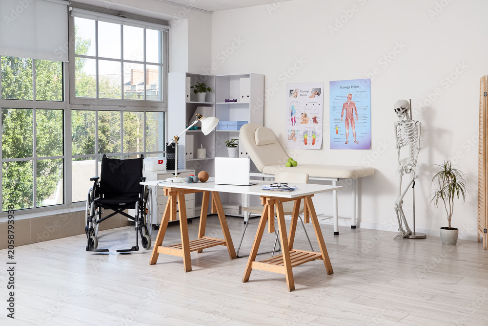 Fototapeta premium Modern physical therapy office interior with wheelchair, examination couch, and anatomical charts for rehabilitation