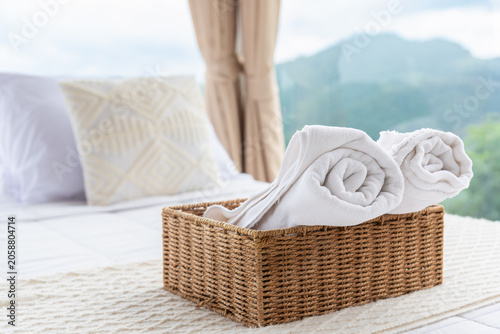White rolled towels in a wicker basket on a bed in a luxury hotel bedroom with a blurry mountain view background.