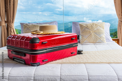 Red suitcase and straw hat on a bed in a luxury hotel room with panoramic glass windows overlooking mountains and a blue sky.