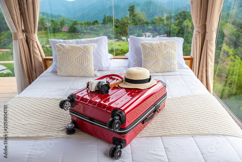 Red suitcase, straw hat, and mirrorless camera on a bed in a luxury hotel room with panoramic glass windows overlooking green mountains.