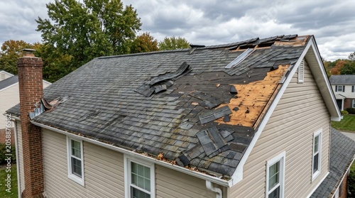 A residential house shows significant damage to its roof. Several shingles are missing, exposing the underlying wood.