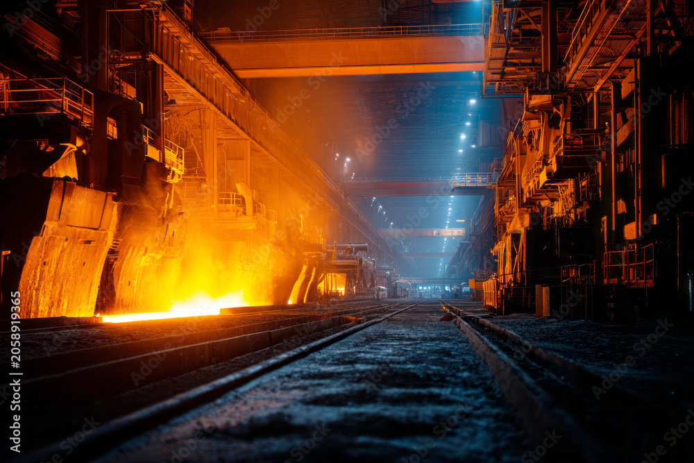 Fototapeta premium Molten steel is pouring from a blast furnace into a channel for casting. The scene shows the interior of a steel mill at night. Bright orange and yellow glow fills the area