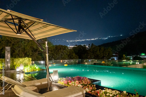 Nighttime landscape from Dehradun featuring a luxury infinity pool, colorful flower beds, and the illuminated Mussoorie hillside
