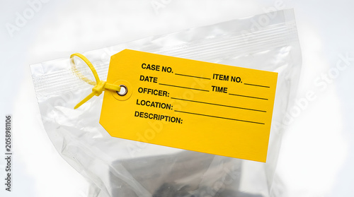 A bright yellow evidence tag, filled with details like case number, date, and officer name, is attached to a clear plastic bag containing an item, all presented in a clean, close-up studio shot.