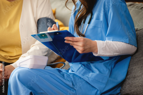 Nurse in blue scrubs checks blood pressure, Caregiver holds clipboard while measures blood pressure with cuff on patient. Patient sits comfortably on couch, relaxed.