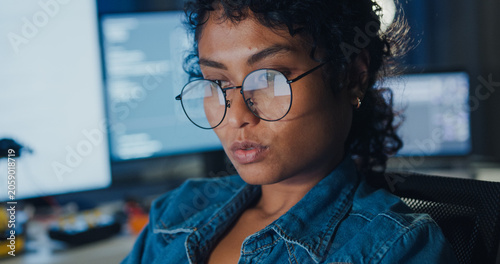 Close-up young Black female programmer coding AI software on multiple screens in night office. Network security, cybersecurity, machine learning, working late, overtime, robotics automation concept.