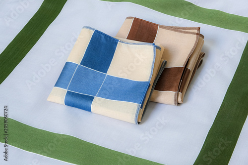 A blue-and-beige and a brown-and-beige folded handkerchiefs neatly arranged together on top of a green-bordered gray cotton handkerchief.