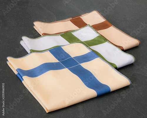 A set of folded cotton handkerchiefs featuring a minimalist cross pattern in blue, green, and brown on a textured dark background.
