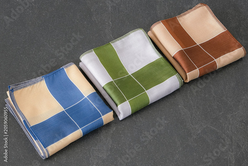 A set of folded cotton handkerchiefs featuring a minimalist cross pattern in blue, green, and brown on a textured dark background.