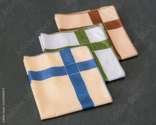 A set of folded cotton handkerchiefs featuring a minimalist cross pattern in blue, green, and brown on a textured dark background.