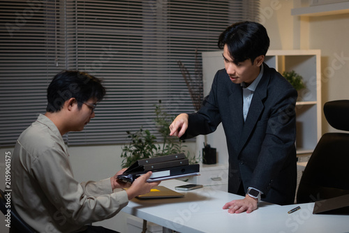 Obraz na płótnie The tense office atmosphere at night: an Asian male supervisor or manager, wearing a business suit, stands leaning over his desk and pointing his finger, giving orders or reprimanding male employees