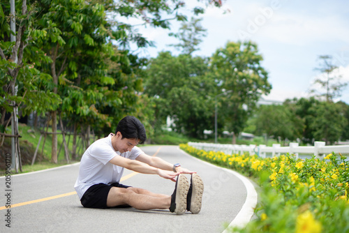 A young man in a white t-shirt and black shorts, dressed in athletic wear, sat on the paved road of the bicycle lane in a park. He was bending over, stretching his leg muscles.