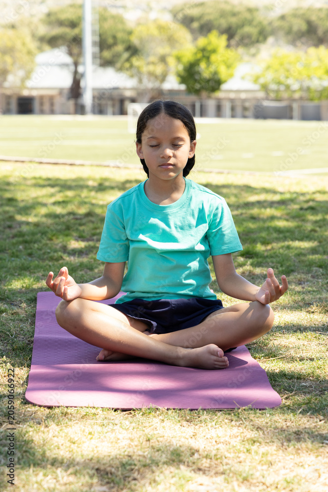 Fototapeta premium Female child sitting on purple yoga mat at park, wearing turquoise tee and navy shorts, meditating