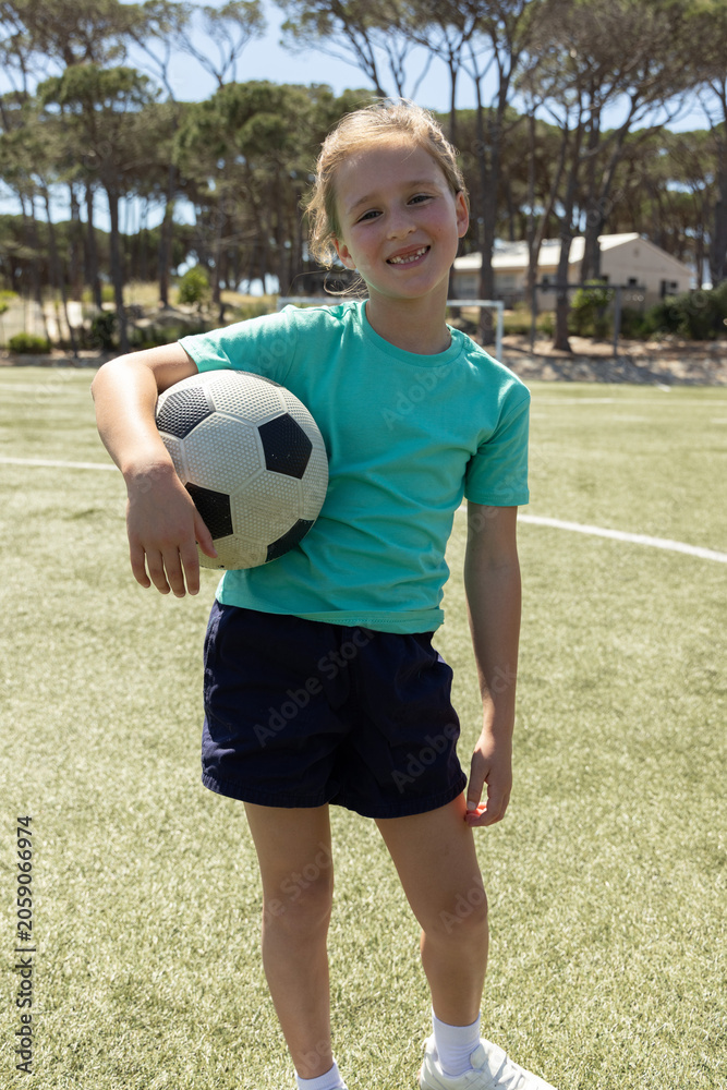 Obraz premium Girl child standing on grass pitch holding black-white soccer ball, wearing teal shirt, navy shorts