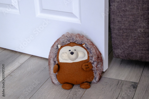 Felt hedgehog door stopper rests beside white door. Soft, round, friendly face invites warmth and humor. Wooden floor and muted tones create cozy, inviting atmosphere. Perfect for homes, stores