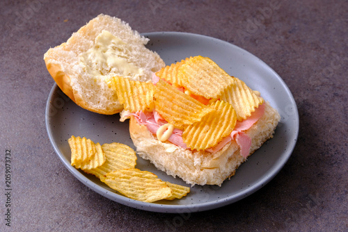 Crunchy chips rest atop a sandwich. Ham and cheese fill soft bread. Halved roll sits beside the sandwich. Gray plate holds the meal. Dark surface enhances visual focus