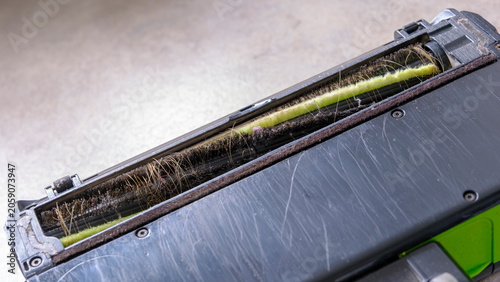 Dirty vacuum brush head shows clogged bristles. Hair and debris fill green interior strands. Mud stains and dust cling to metal casing. Tool looks worn, used for heavy cleaning