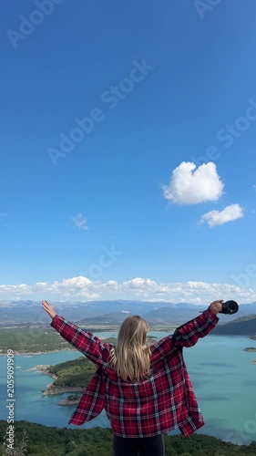 Young blonde woman walking with open arms and holding a camera while enjoying a panoramic view of Vilusi Ponikvica lake, blue sky, mountains and nature in Montenegro