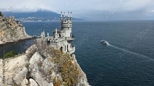 Swallow's nest castle, a neo-gothic landmark, standing perched on a dramatic rock cliff above the dark black sea, with a tour boat navigating the water creating a wake