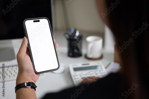 A woman holds a smartphone with a blank white mockup screen over a busy office desk.