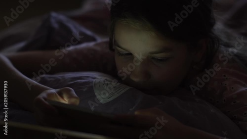 A teenage girl lies in bed before sleep and holds her phone in her hands scrolling through her online feed, which illuminates her face. High quality FullHD footage