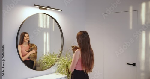 This stylish interior features a round mirror, elegant lighting, and lush greenery for a chic ambiance. A woman cradles cats by a sunlit window, sharing a serene moment