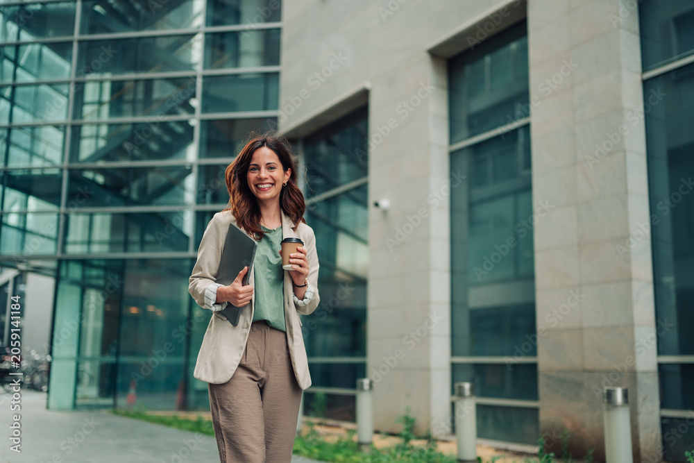 Fototapeta premium Professional businesswoman smiling, walking confidently with laptop and coffee, ready for work in an urban office area