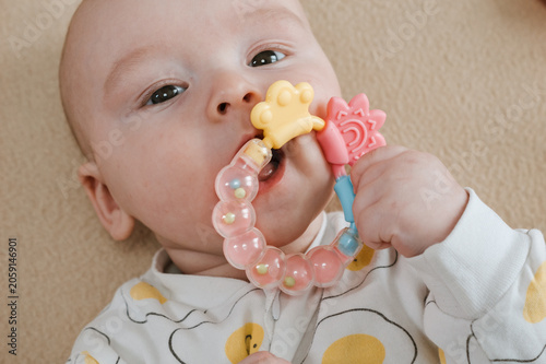 Baby lying on bed and gripping teething ring with both hands. Child chewing toy for teething relief. Exploring textures while soothing sore gums. Sensory discovery at home.