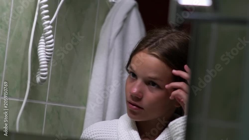 A teenage girl in a hotel bathroom, looking in the mirror, adjusting her long hair. A girl in a bathrobe looks in the mirror. High quality FullHD footage