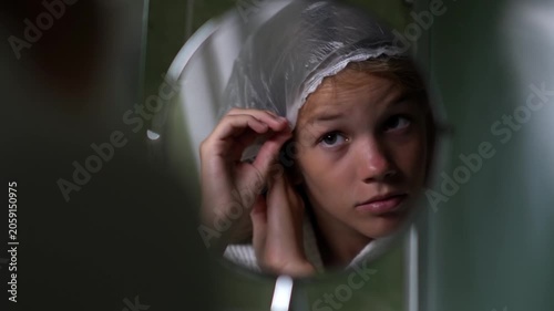 A girl looks at herself in a round mirror in a hotel bathroom and adjusts the clear shower cap on her hair. The teenager is performing her morning or evening body care routine.