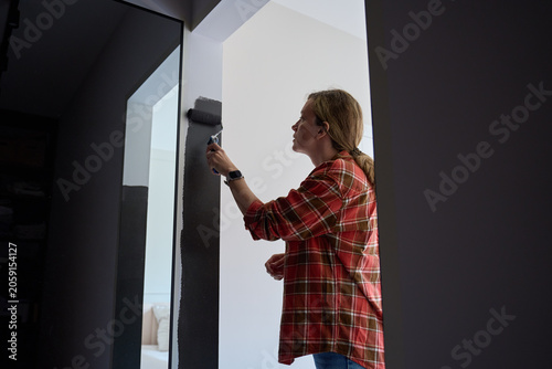 Woman applies dark paint to wall with roller near mirror and white doorway