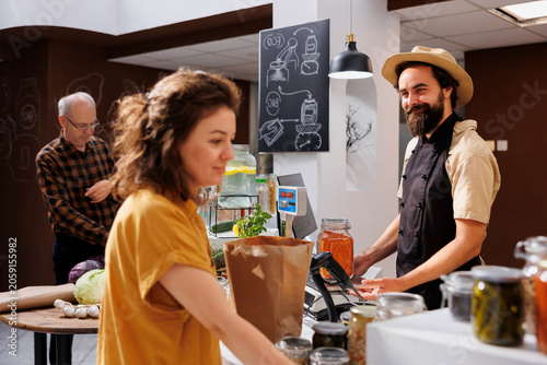 Картината върху платно Cheerful retailer at checkout counter in zero waste shop weighting vegan products purchased by shopper, woman purchasing healthy food in local neighborhood store with biodegradable paper bags