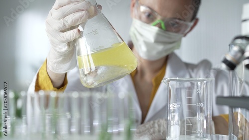 Cuadro en lienzo Woman scientist researcher wearing white protective gloves is shaking up a yellow chemical solution inside an Erlenmeyer flask in a laboratory setting, portrait view
