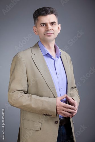 Portrait of Confident Positive Mature Gray-Haired Brunette Caucasian Man 50 Years Old Wearing Pale Jacket and Purple Violet Shirt Looking Straight With Closed Hands Over Gray