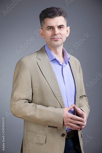 Confident Positive Mature Gray-Haired Brunette Caucasian Man 50 Years Old Wearing Pale Jacket and Purple Violet Shirt Looking Straight With Closed Hands Over Gray Background