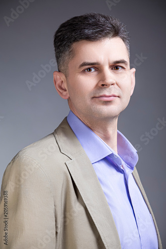 Closeup Portrait of Confident Positive Mature Caucasian Man 50 Years Old Wearing Pale Jacket Looking Straight With Hands in Pockets Over Gray