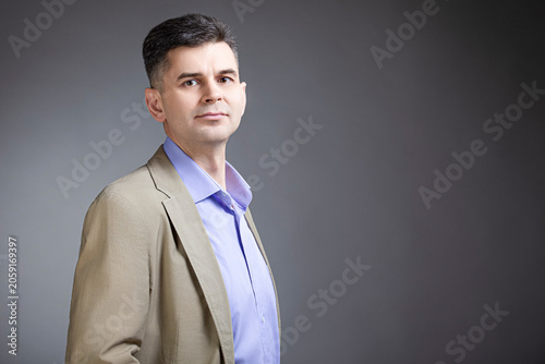 Portrait of Confident Positive Mature Caucasian Man 50 Years Old Wearing Pale Jacket Looking Straight With Hands in Pockets Over Gray