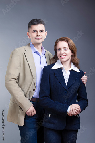 Positive Caucasian Mature Loving Couple Posing in Business Suits Together Embraced Over Gray Backround As Family People in Business Concepts.