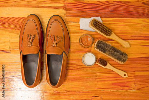 One Pair of Light Tan Tassel Loafers Shoes Made of Calf Leather with Leather Sole Placed In Line With Shoe Cleaning Brushes On Wooden Surface