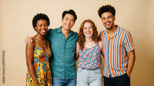 Group of four ethnically diverse young friends smiling and standing together in casual summer clothing against a beige background.