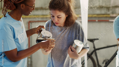Foto African American female volunteer serving a cup of coffee to homeless caucasian lady, friendly black woman at outdoor food bank sharing nourishments to the needy and less fortunate