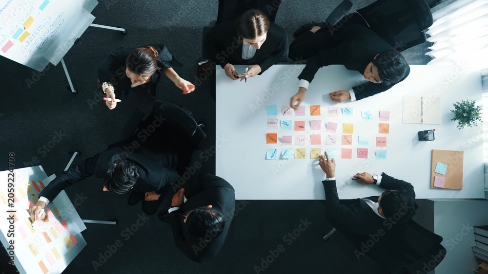 Obraz premium Top down aerial view of business group sharing and brainstorming idea by using sticky notes at meeting room. Investor writing marketing strategy on board while receive note from manager. Directorate.