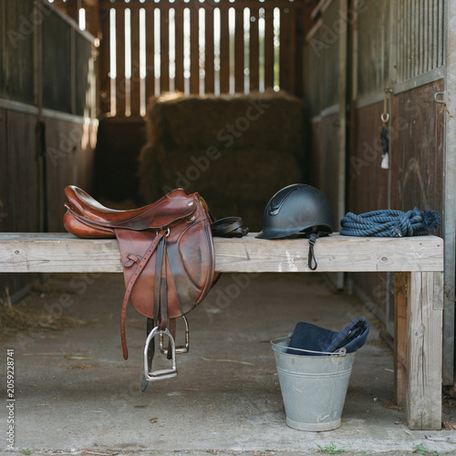 Horse saddle and riding helmet rest upon a rustic wooden bench inside a stable with hay in the background.