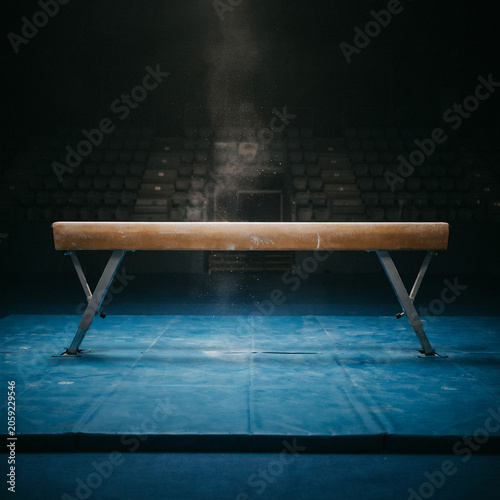 Gymnastics balance beam rests silently on a blue mat within a dark and moody training hall during practice time.